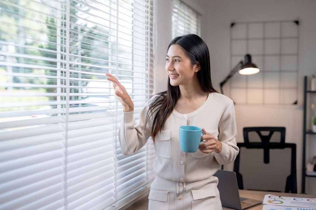 A smiling woman peers through window blinds while drinking her morning coffee